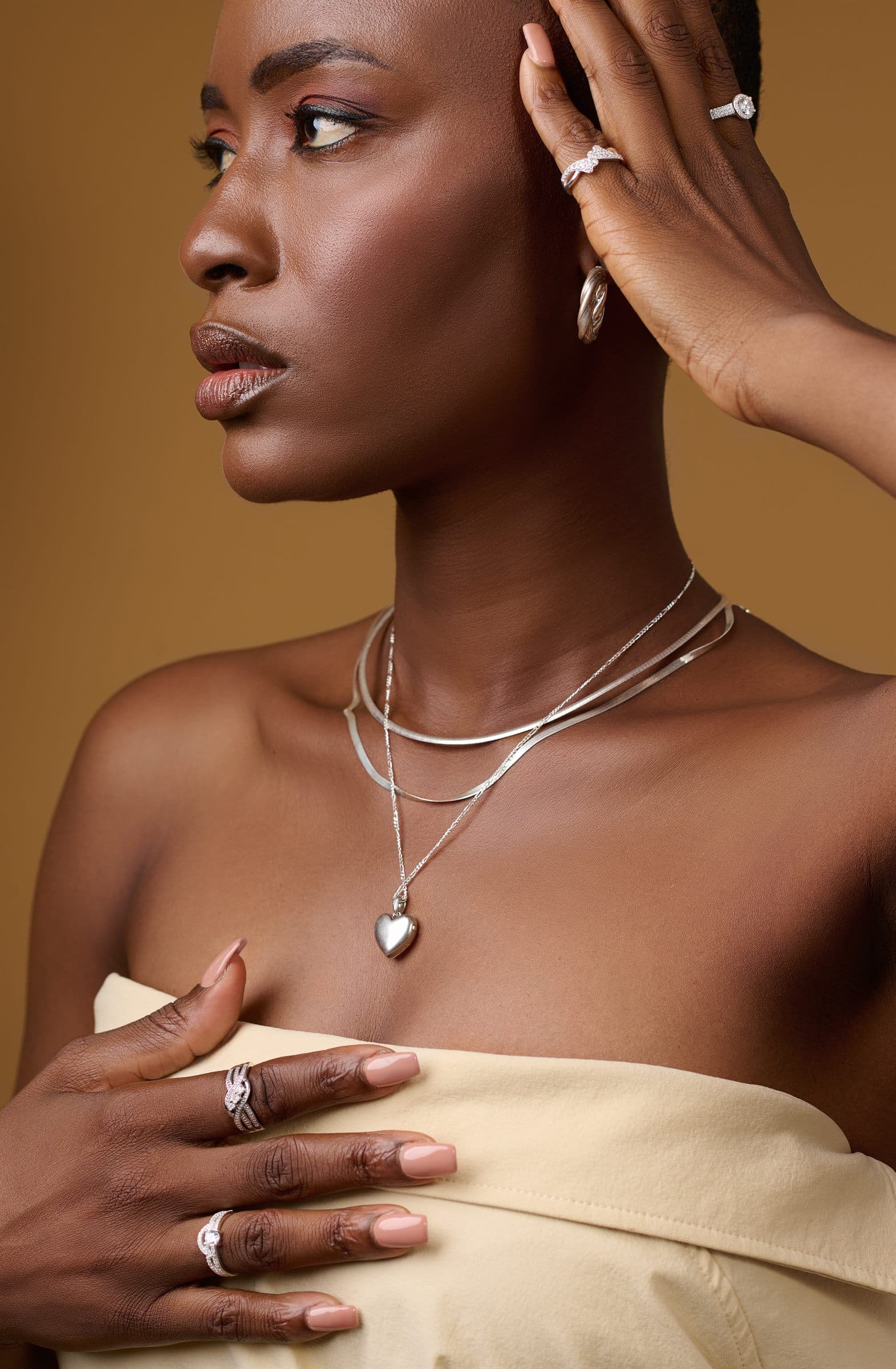 Portrait with a raised hand, layered necklaces against a tan backdrop