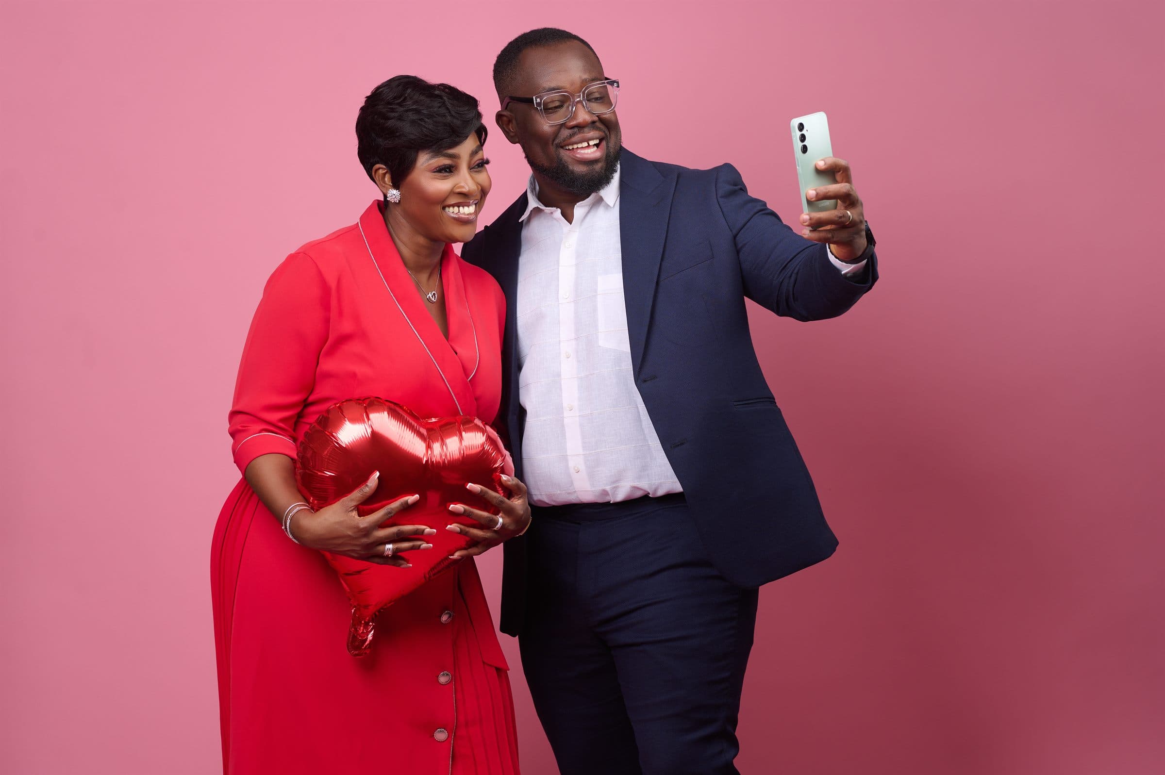 Couple on a pink backdrop with a heart balloon, taking a selfie